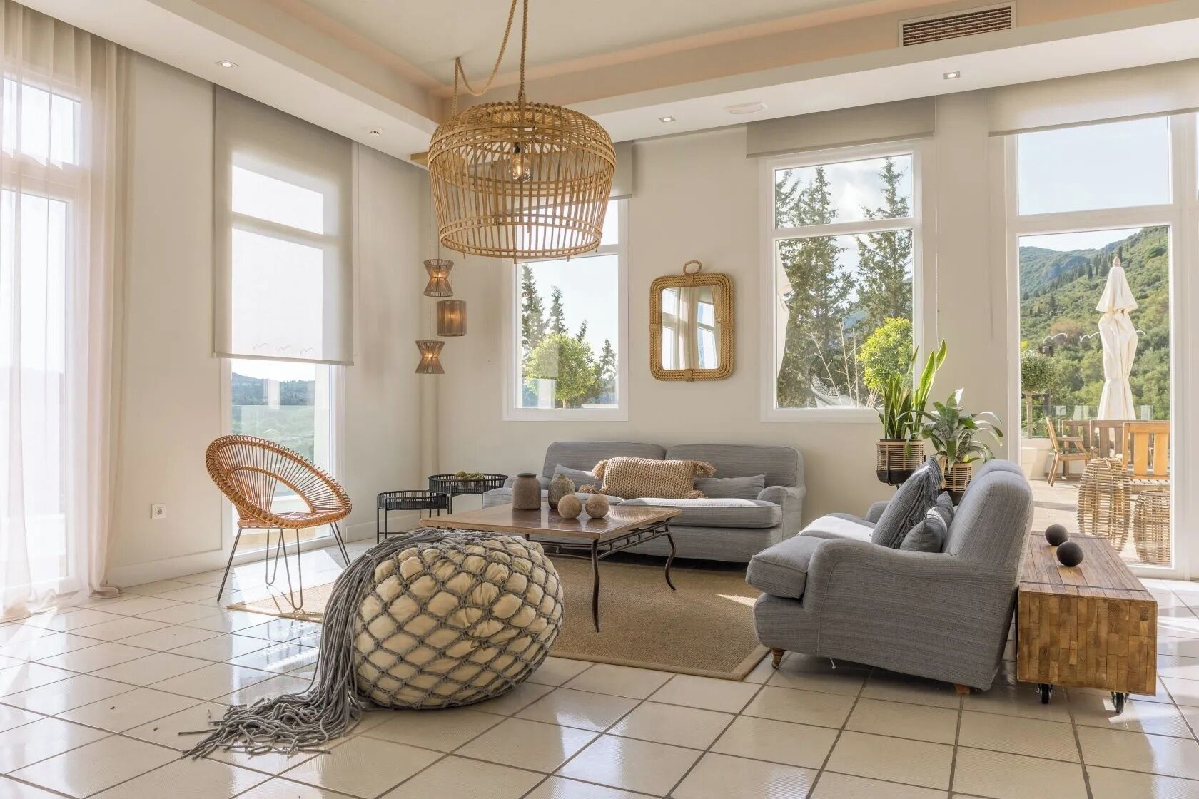 Ein geräumiges Wohnzimmer mit grauem Sofa, Korbstühlen, einem großen gewebten Pouf und Blick ins Grüne durch große Fenster.
