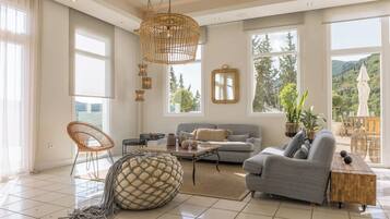 Ein geräumiges Wohnzimmer mit grauem Sofa, Korbstühlen, einem großen gewebten Pouf und Blick ins Grüne durch große Fenster.