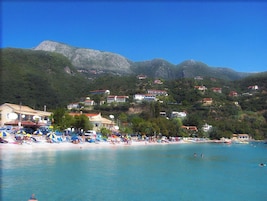 Zona costera con playa, casas en una ladera y agua azul cristalina.