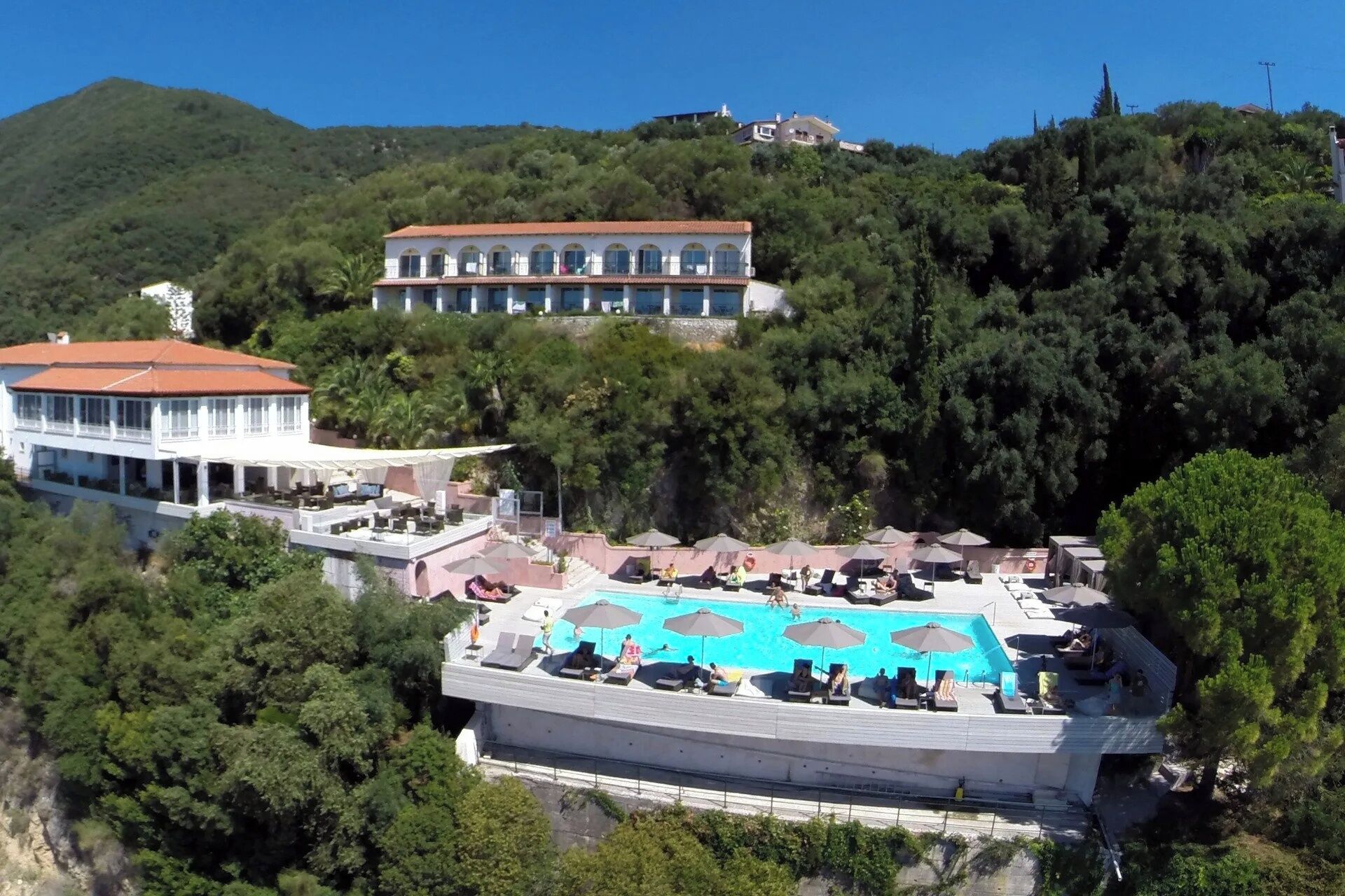 Un complexe hôtelier doté d’une piscine, d’un bâtiment blanc avec un toit rouge, et d’une colline parsemée d’arbres, avec un autre bâtiment visible au loin.