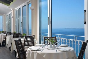Ein Restaurant mit gedeckten Tischen, großen Fenstern und Meerblick.