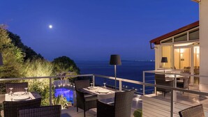 A terrace with dining furniture, a pool, and a view of the sea at night. - Marilena Sea View (Pirgi)