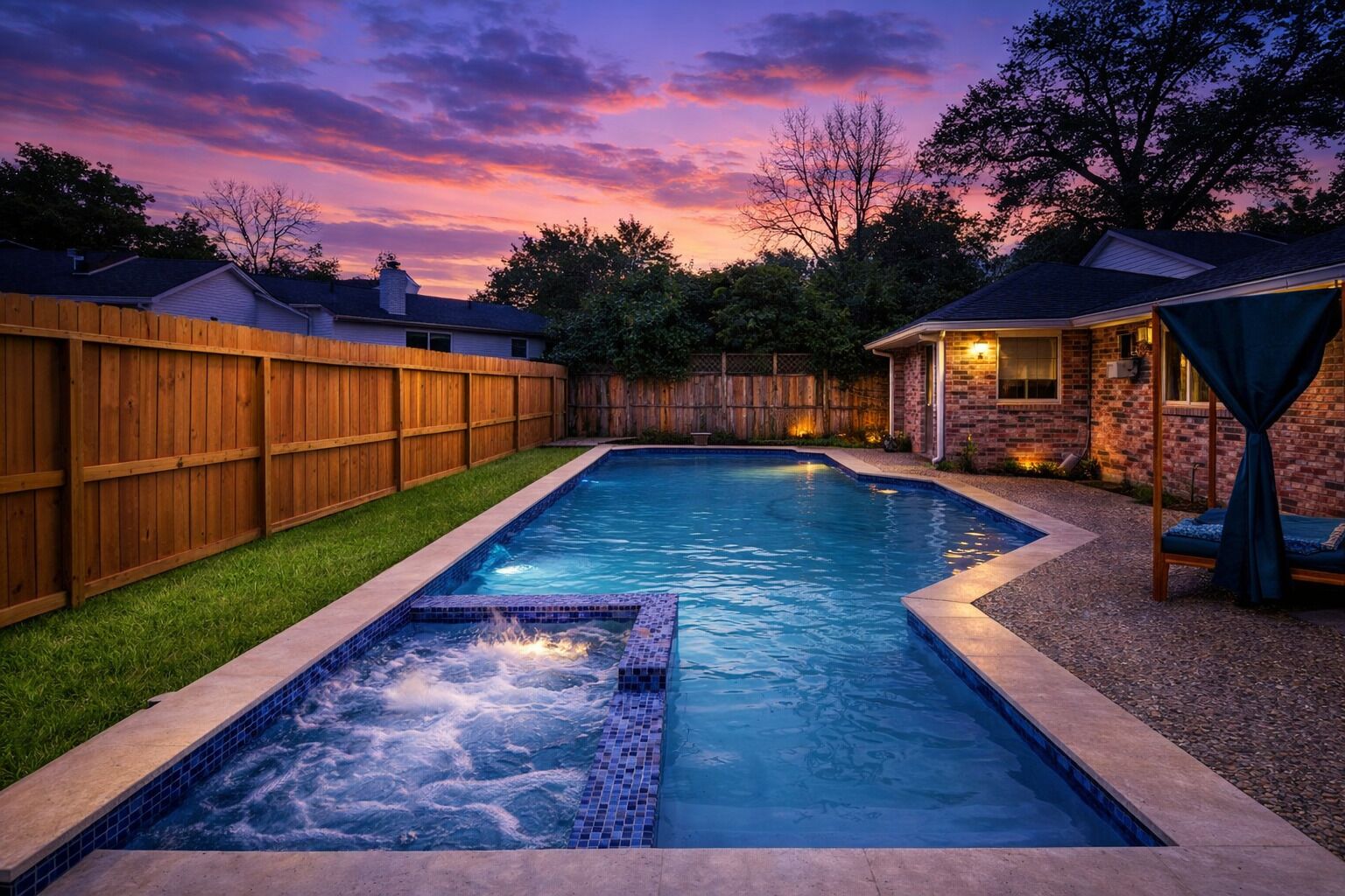A rectangular swimming pool with a hot tub, surrounded by a wooden fence and a brick house with outdoor lighting.