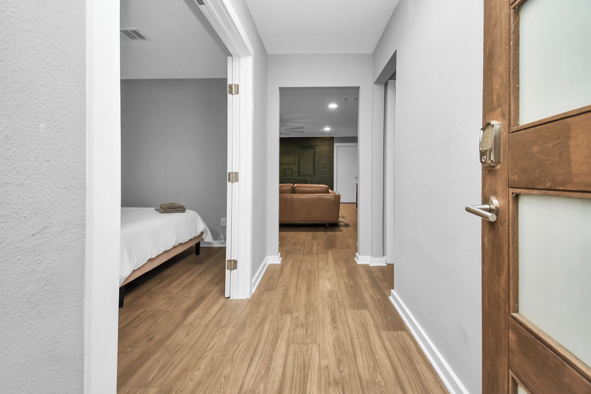 A narrow hallway with a bed on the left, a wooden door on the right, and a glimpse of a room with a brown sofa in the distance.