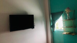 Una habitación con una televisión de pantalla plana en la pared, una ventana con cortinas y una estantería con un jarrón y objetos decorativos.