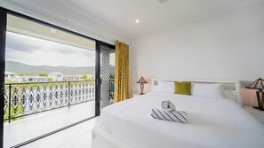 Quarto de hotel com cama, mesa de cabeceira, luminária, janela com vista para casas e montanhas, e varanda com guarda-corpo.