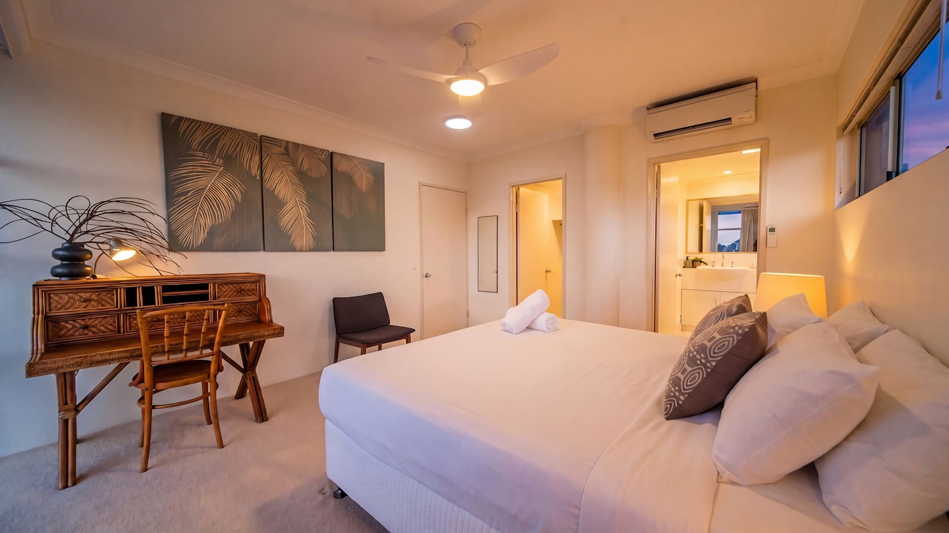 A hotel room with a bed, a desk, a chair, a ceiling fan, and a large window.