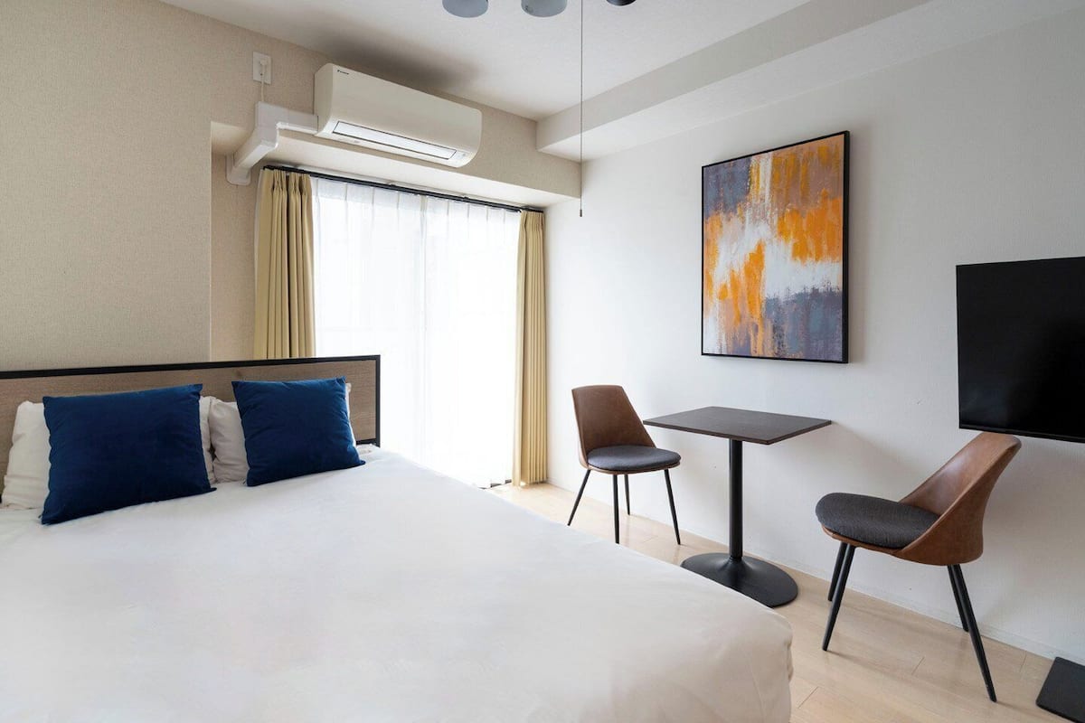 Habitación de hotel con cama, dos sillas, una mesita, aire acondicionado, un cuadro y televisor.