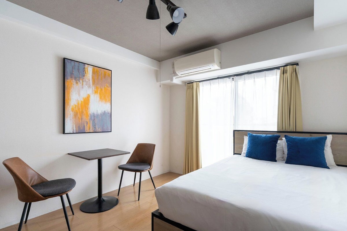 Habitación de hotel con cama, dos sillas, una mesa, aire acondicionado y ventana con cortinas.
