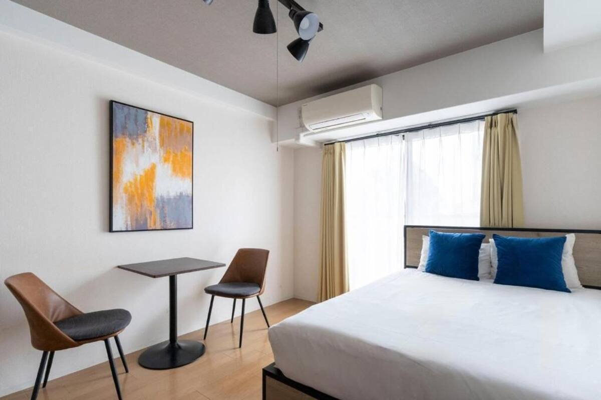 Habitación de hotel con cama, dos sillas, una mesa, aire acondicionado y ventana con cortinas.