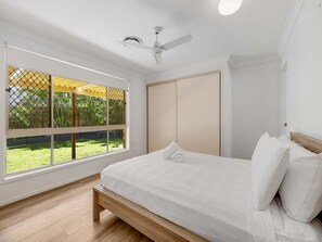 Ein Schlafzimmer mit Bett, Deckenventilator, Fenster mit Blick auf Grünflächen und einer Schiebetür.