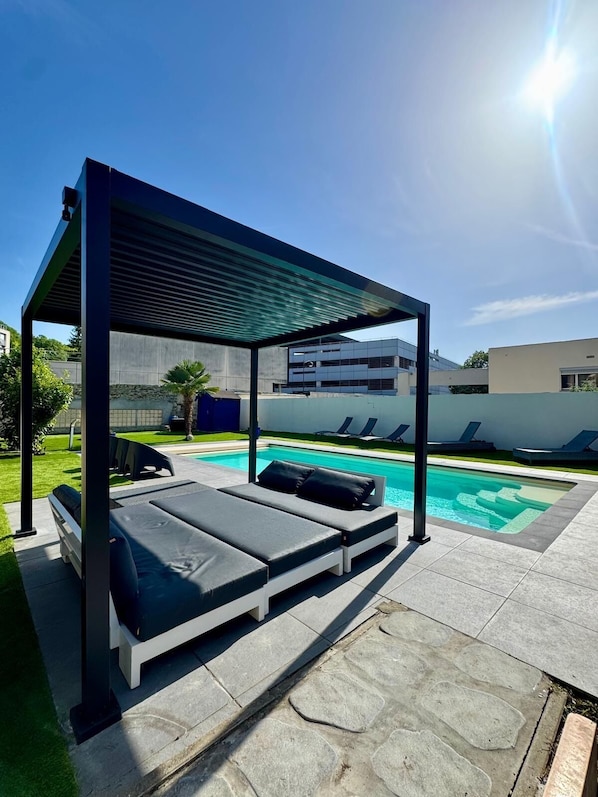 Un área de piscina moderna al aire libre con una zona de estar cubierta y sillones reclinables.