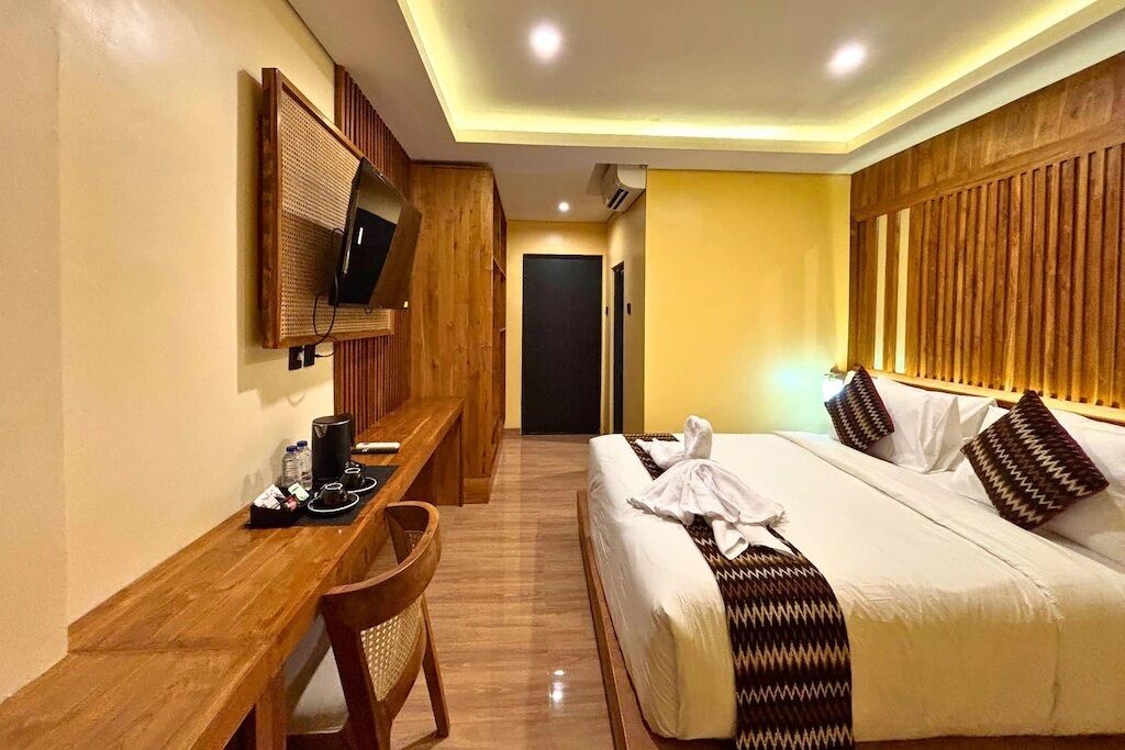 Ein Hotelzimmer mit Holztisch, einem Bett mit gemusterten Kissen, einem an der Wand befestigten Fernseher und einer Tür im Hintergrund.
