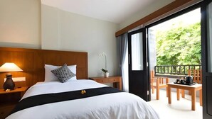 Habitación de hotel con cama, mesita de noche, lámpara, planta en maceta y balcón con mobiliario de madera.