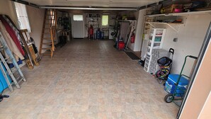 A garage with a tiled floor, a ladder, and various items including a blue surfboard, a red fire extinguisher, and a green cart. - SEPTEMBER SPECIAL: Vacation Rental in St Gilles Croix de Vie (Le Fenouiller)