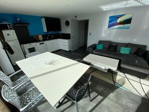 A modern kitchen with white cabinets, a black countertop, and a built-in oven. There is a dining area with a white table and chairs, and a gray sofa with teal cushions. - Le Corbier lodge 43 2-bedroom apartment at the foot of the slopes (Villarembert)