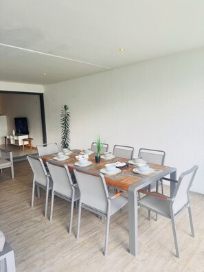A dining area with a long table set for six, surrounded by chairs, with a potted plant on the table and a television in the background. - Latitude Luxury Seafront Apt (Grande Riviere Noire)
