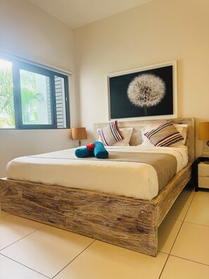 A neatly made bed with a wooden headboard, a large window with blinds, a framed artwork above the bed, and a bedside table with a lamp. - Latitude Luxury Seafront Apt (Grande Riviere Noire)