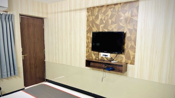 A hotel room with a bed, a flat-screen TV mounted on a wooden panel wall, a wooden bedside table, and a door. - Rosee Wood (Visakhapatnam)
