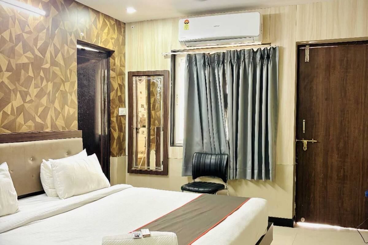 Habitación de hotel con cama, ventilador de techo, aire acondicionado, espejo, silla y puerta.