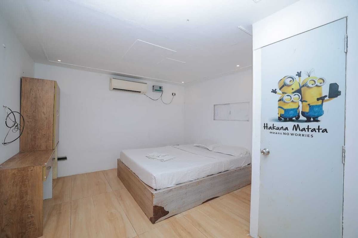Un dormitorio con estructura de cama de madera, pared blanca, unidad de aire acondicionado y una puerta con un sticker de Minions.