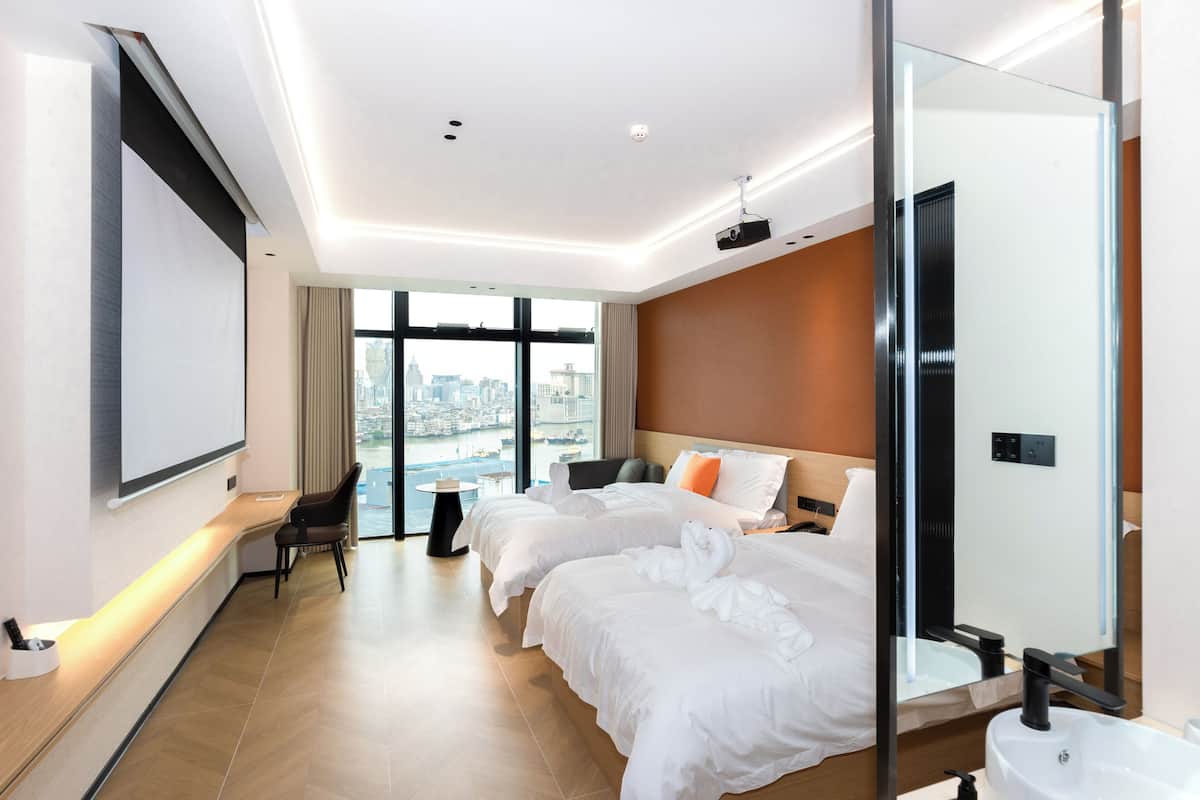 Une chambre d’hôtel avec deux lits, un bureau, une chaise, une grande fenêtre donnant sur la ville et une salle de bain moderne.
