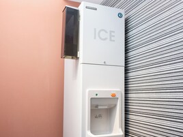 Een witte ijsdispenser met een drukknoop en een indicatorlampje aan een muur.