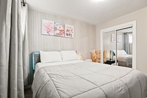 A bedroom with a bed, a bedside table, a mirror, and a wall with vertical lines and floral artwork. - Cozy 1-Bedroom Apartment in Calgary with Kitchenette (Calgary)