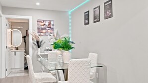 Uma área de refeições moderna com uma mesa de vidro, cadeiras brancas, um vaso com plantas e cartazes motivacionais emoldurados na parede.