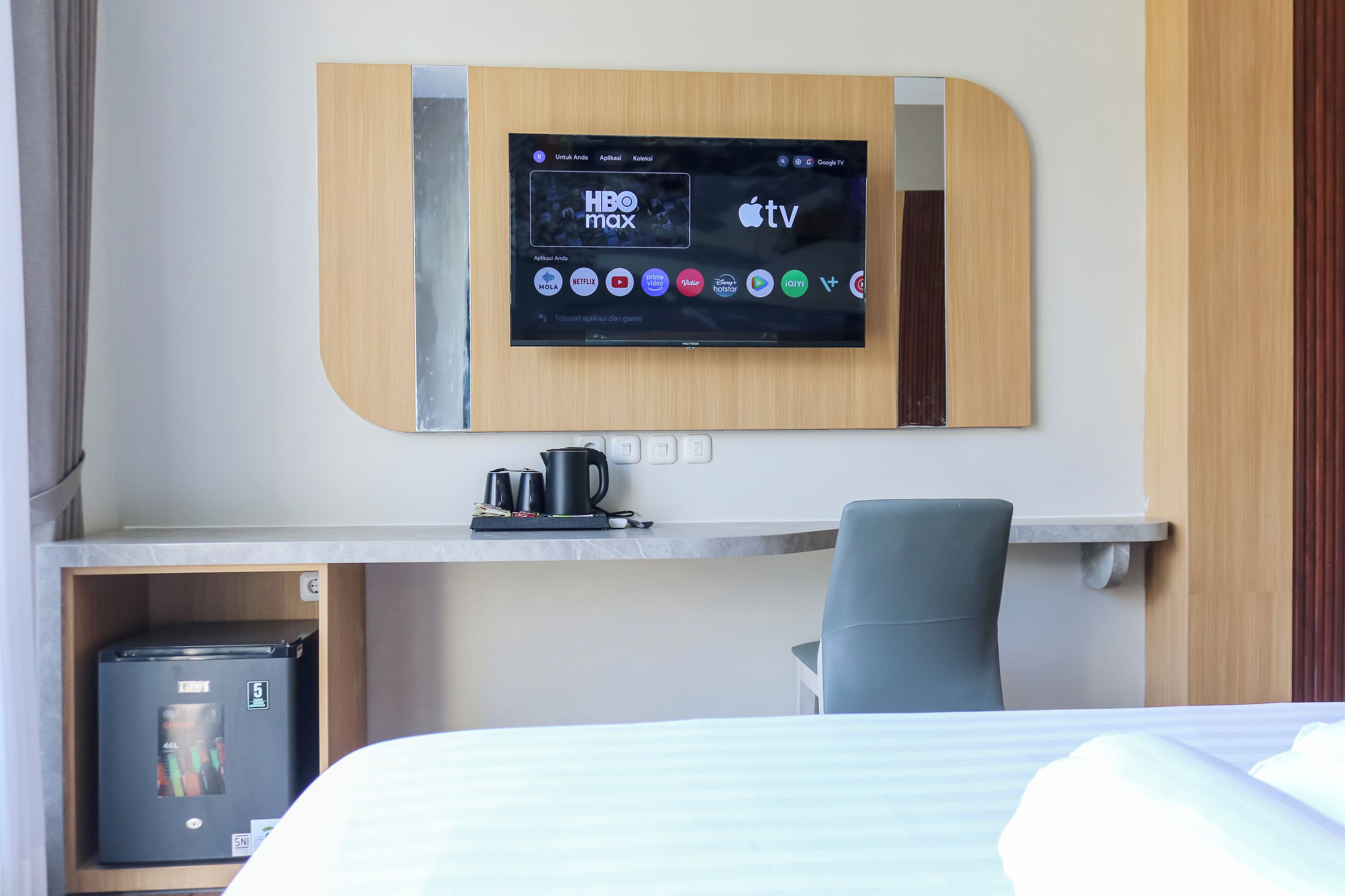 Una camera d'albergo con TV a parete che mostra HBO Max e Apple TV, una scrivania con una macchina per caffè e un frigorifero compatto.