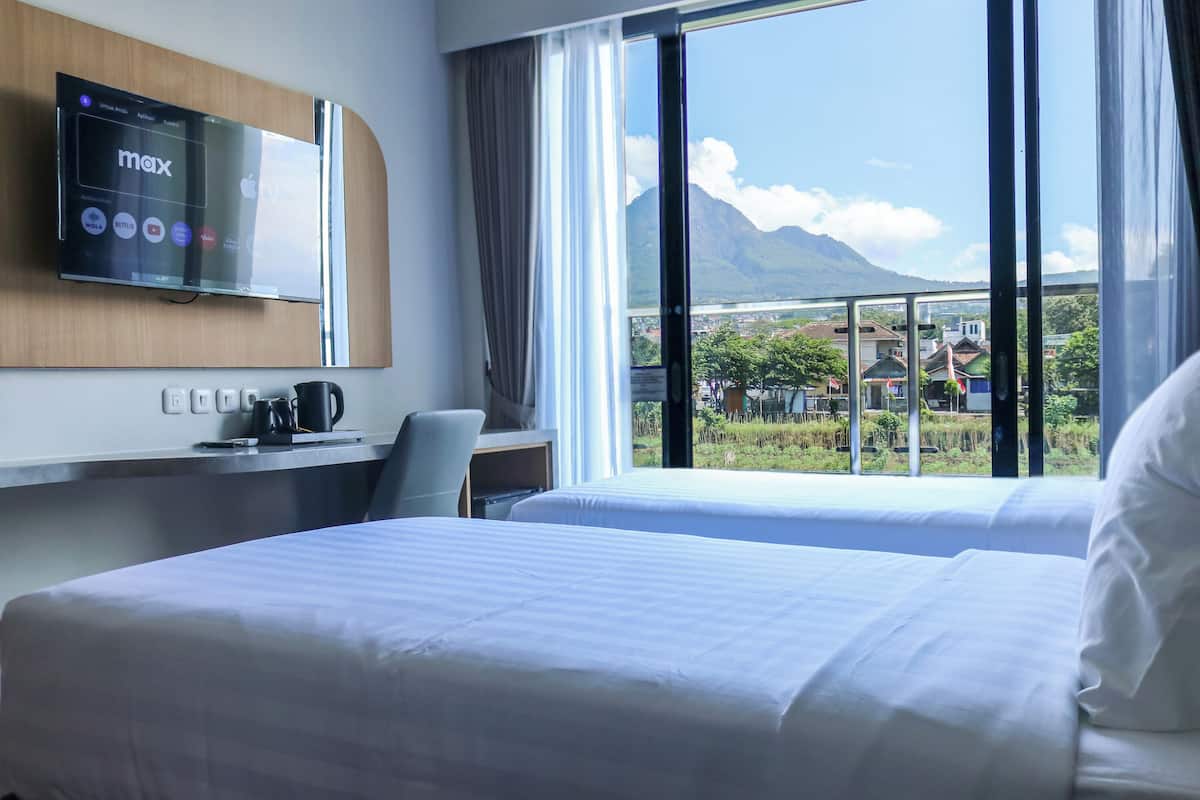 Una habitación de hotel con cama, escritorio con cafetera, televisor montado en la pared y ventana grande con vistas a montañas y edificios.