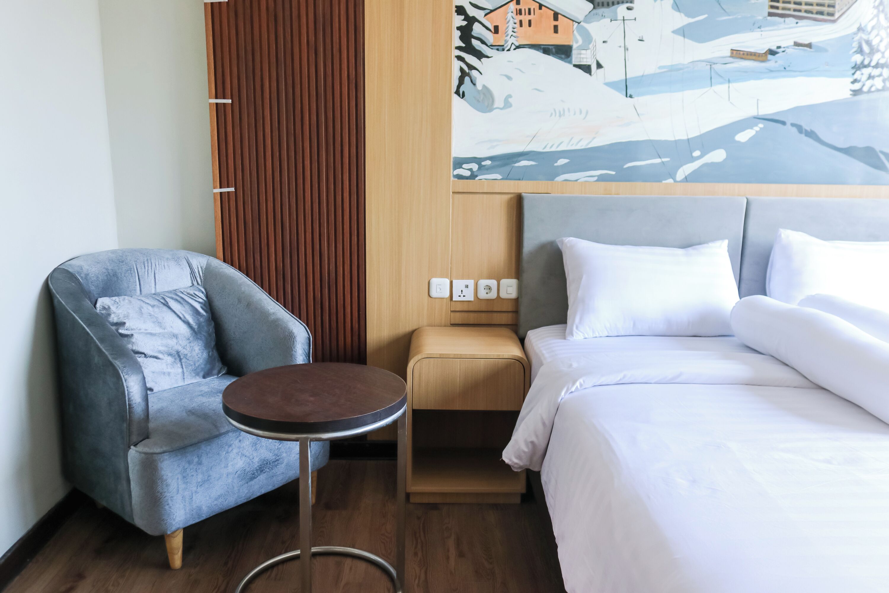 Een hotelkamer met een bed, een grijze fauteuil, een klein rond tafeltje, een houten nachtkastje en een stopcontact op de muur.
