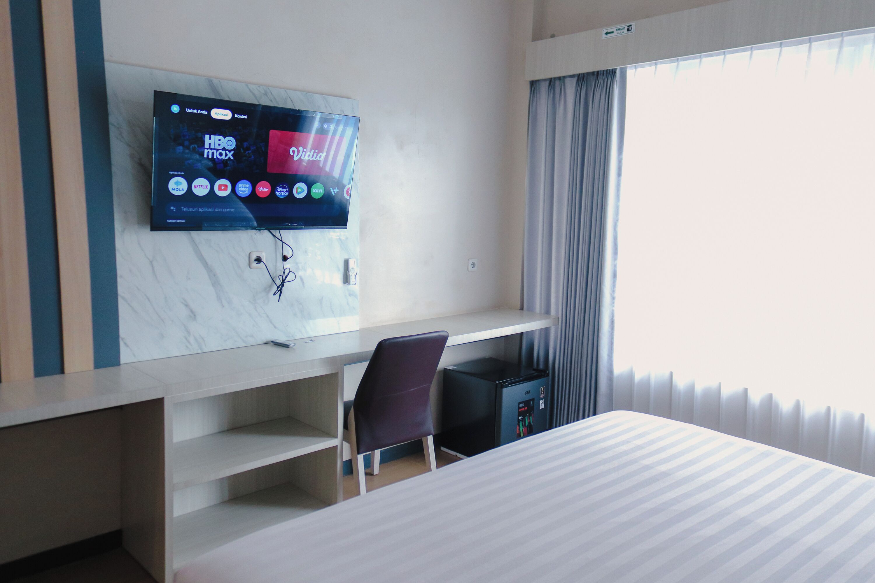 Habitación de hotel con televisión mostrando servicios de streaming, una cama, un escritorio con silla y una ventana con cortinas.
