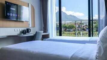 Habitación de hotel con una cama, un escritorio con cafetera, un televisor en la pared y un ventanal con vistas a montañas y edificios.
