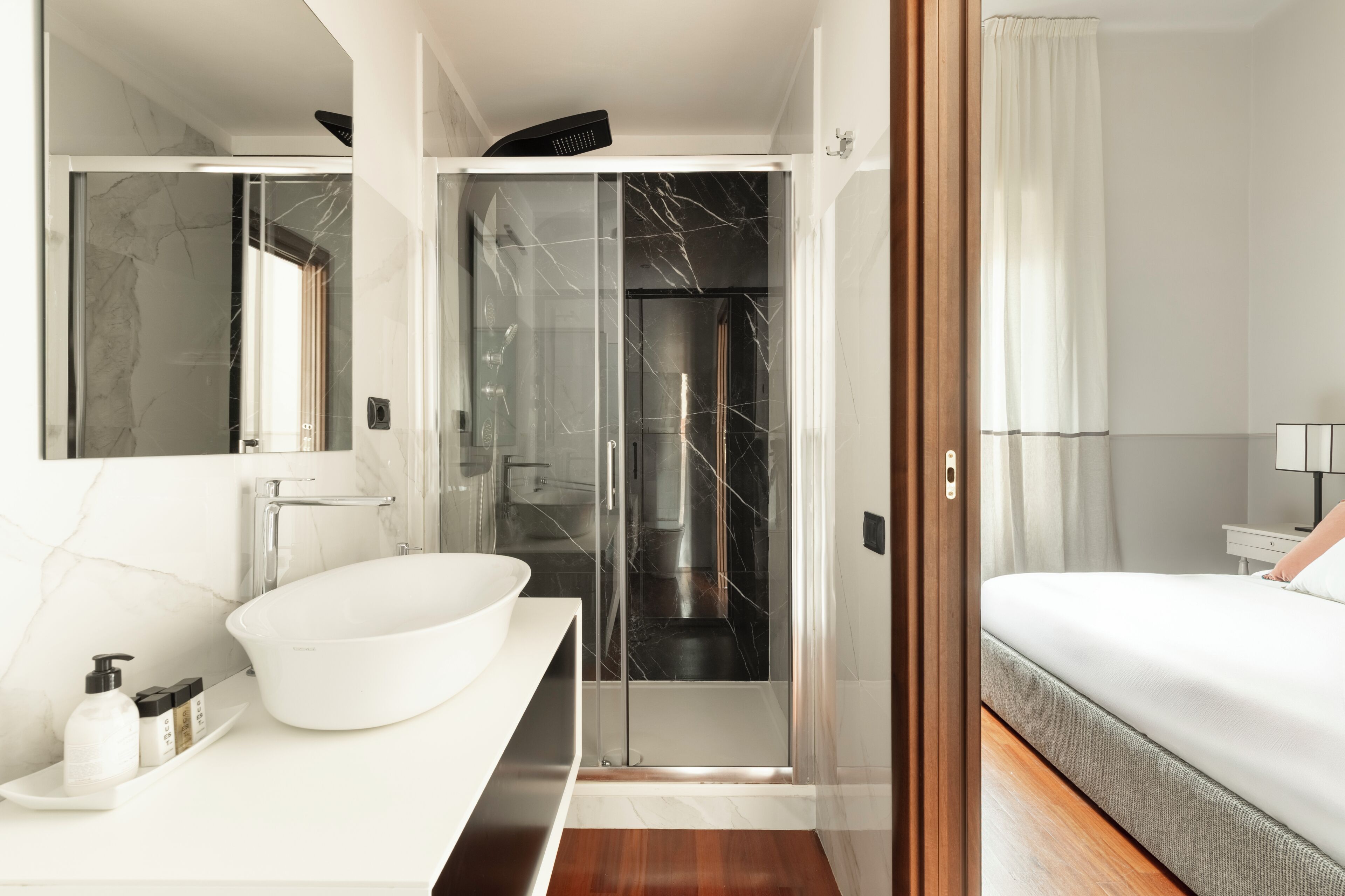 A modern bathroom with a large white sink, a glass shower enclosure, and a bed with a white comforter.