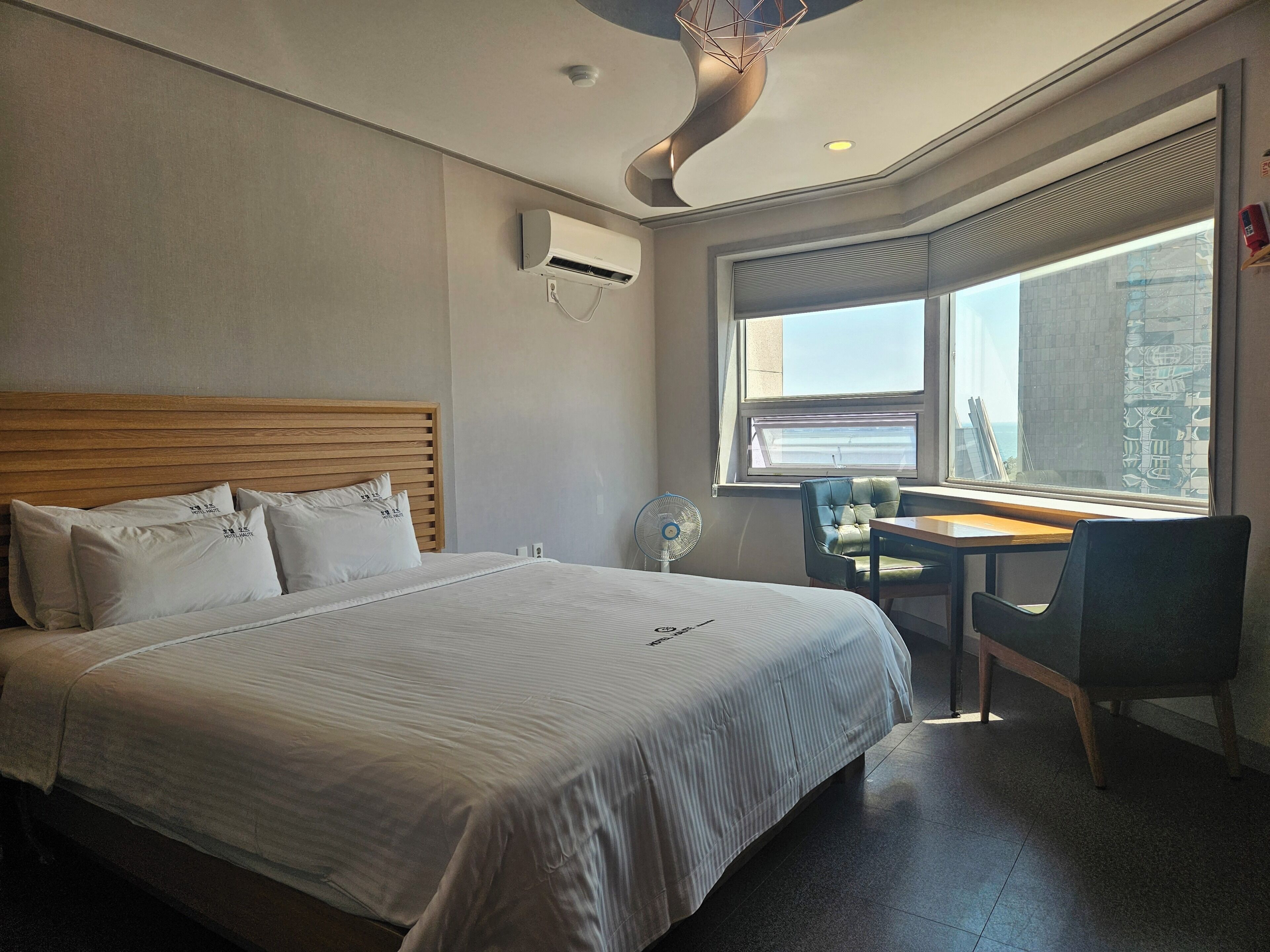 A hotel room with a bed, a desk, a chair, a window with a view, and an air conditioning unit.