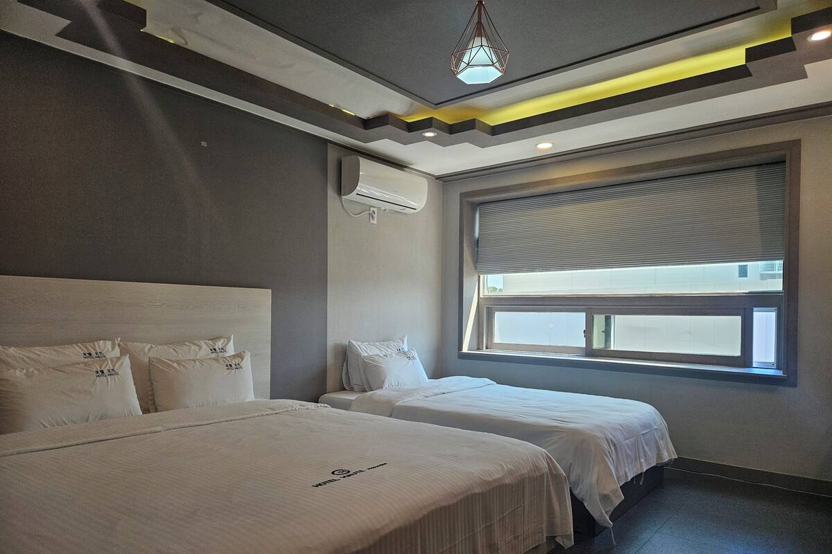 A hotel room with two beds, a large window with blinds, an air conditioning unit, and a hanging light fixture.