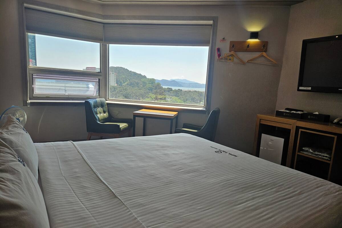 A hotel room with a large bed, a desk, a chair, a TV, and a window with a view of a mountain and cityscape.