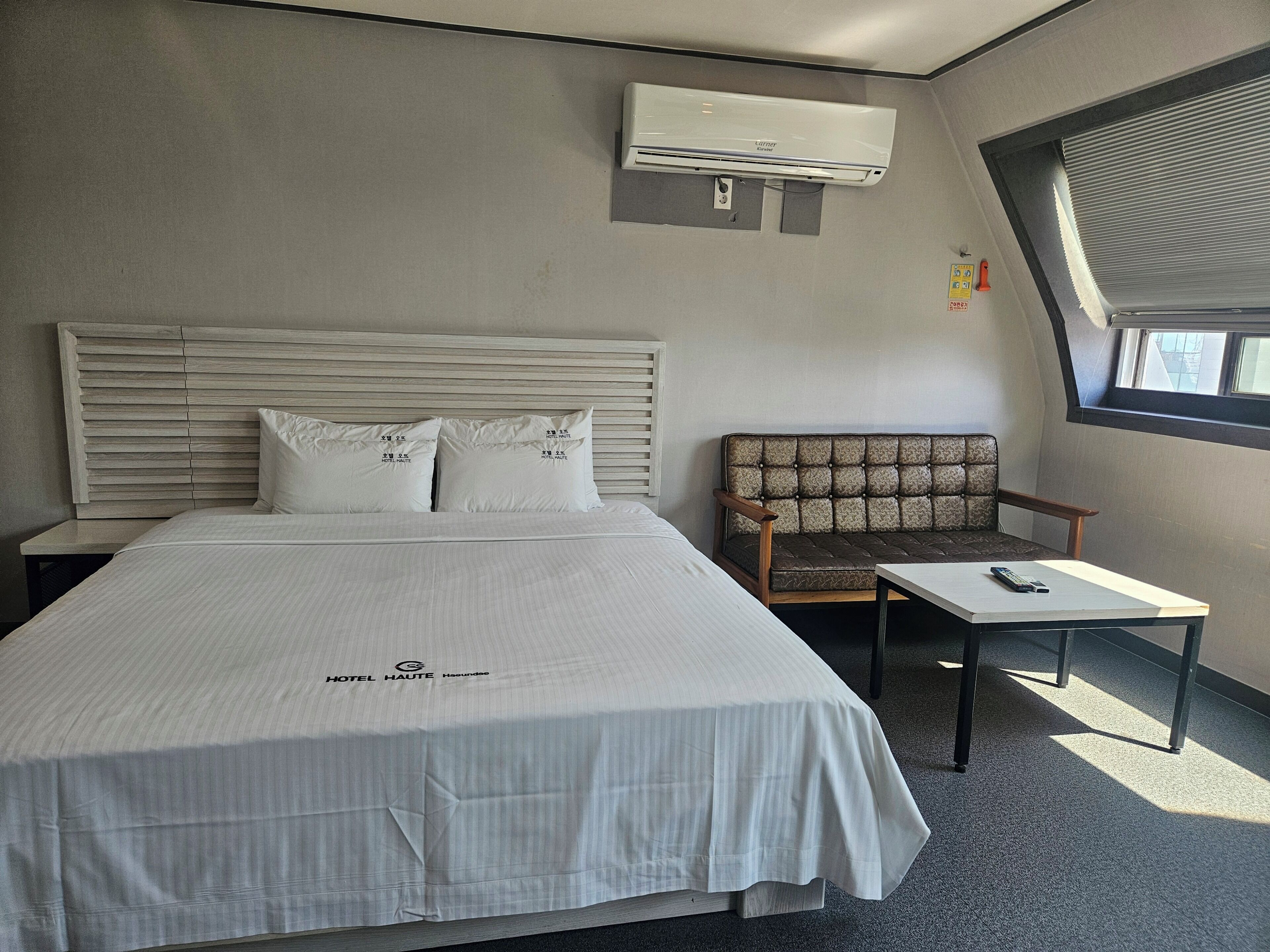 Une chambre d’hôtel avec un lit, une chaise, une petite table, un appareil de climatisation et une fenêtre.