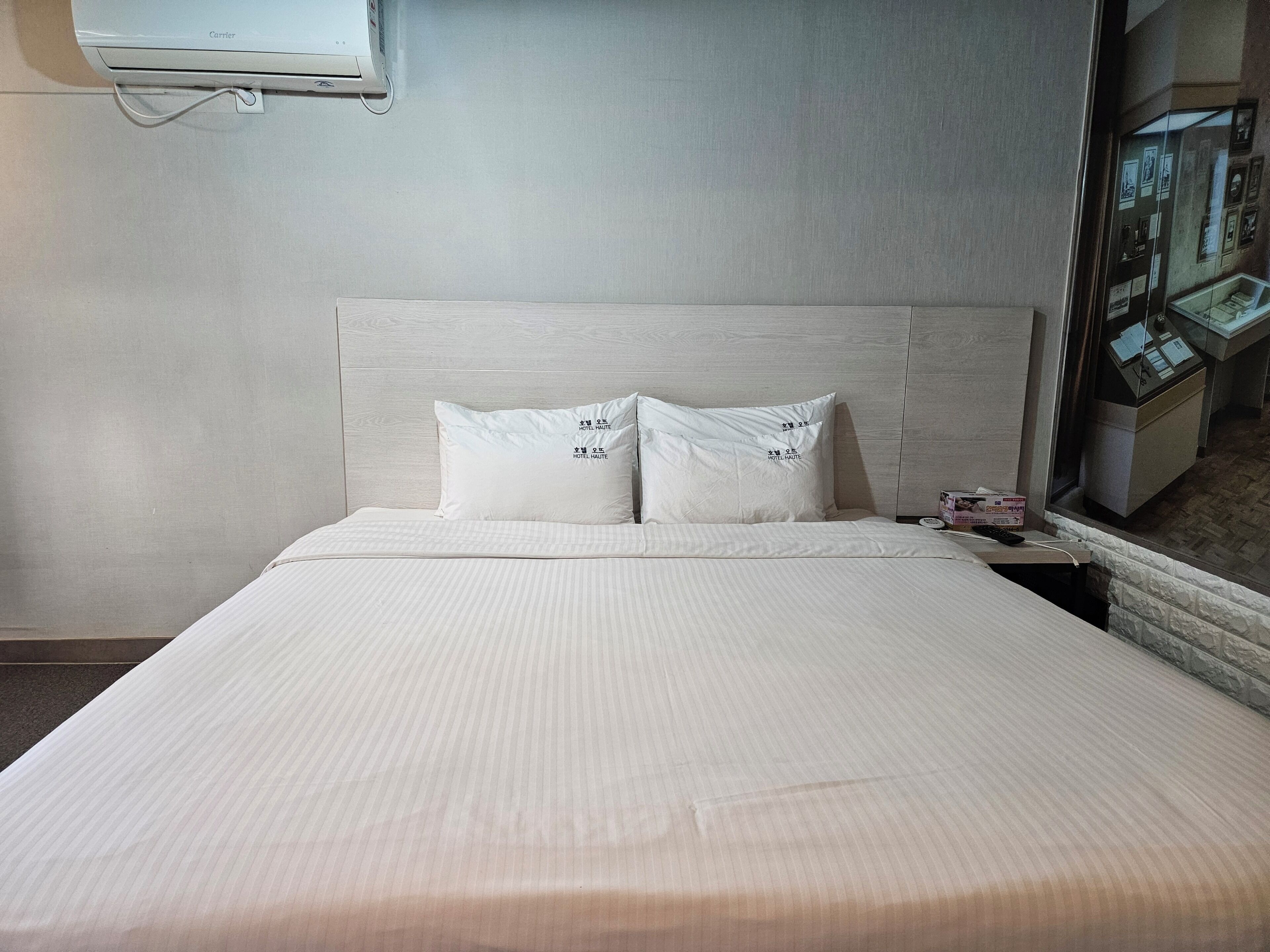 Ett hotellrum med en säng, två kuddar, ett fönster med utsikt över lobbyn och en väggmonterad luftkonditioneringsanläggning.