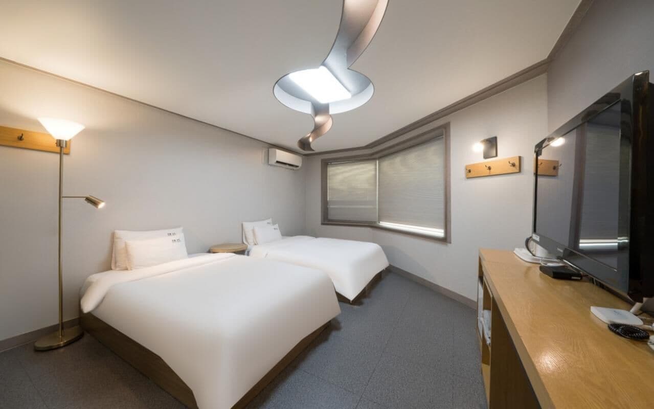A hotel room with two beds, a desk with a flat-screen TV, a large mirror, and a ceiling light fixture.