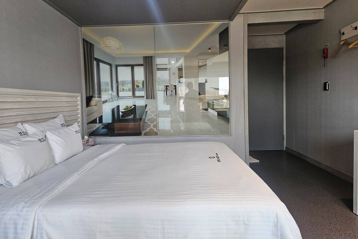 A modern hotel room with a large bed, a glass wall, and a view of the dining area.
