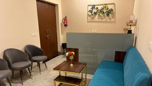 A living room with a blue sofa, two grey armchairs, a small wooden coffee table, and a decorative wall piece. - Hotel Highway Retreat (Dhanaura)