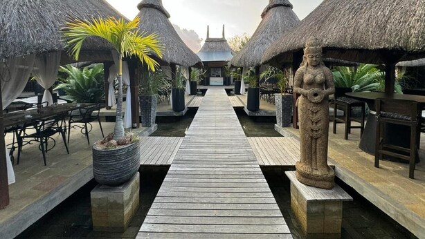 A wooden pathway leading to thatched-roof structures with outdoor seating and a statue.