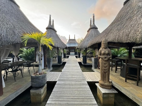 A wooden pathway leading to thatched-roof structures with outdoor seating and a statue.