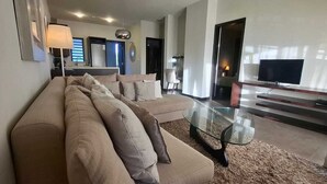 A modern living room with a sofa, a glass coffee table, a flat-screen TV, and a kitchen area in the background.