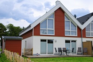 A two-story house with a red and white exterior, large windows, and a patio with outdoor furniture. - Vacation home Blinkfüer in Nordhorn, modern furnished vacation home with 86m² living space on 2 floors. With whirlpool, sauna and fireplace. Sunny terrace with garden furniture and parasol. Bicycle shed (Nordhorn)