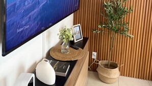Ein flachbildfernseher an der Wand, eine Wand verkleidet mit Holzpaneelen, ein gerahmtes Bild, eine kleine Topfpflanze und ein weißer Lautsprecher.