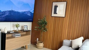 Ein modernes Wohnzimmer mit einem großen Flachbildfernseher, einer Wand mit Holzpaneelen, einem grauen Sofa, einem kleinen Beistelltisch und einem Blumentopf.