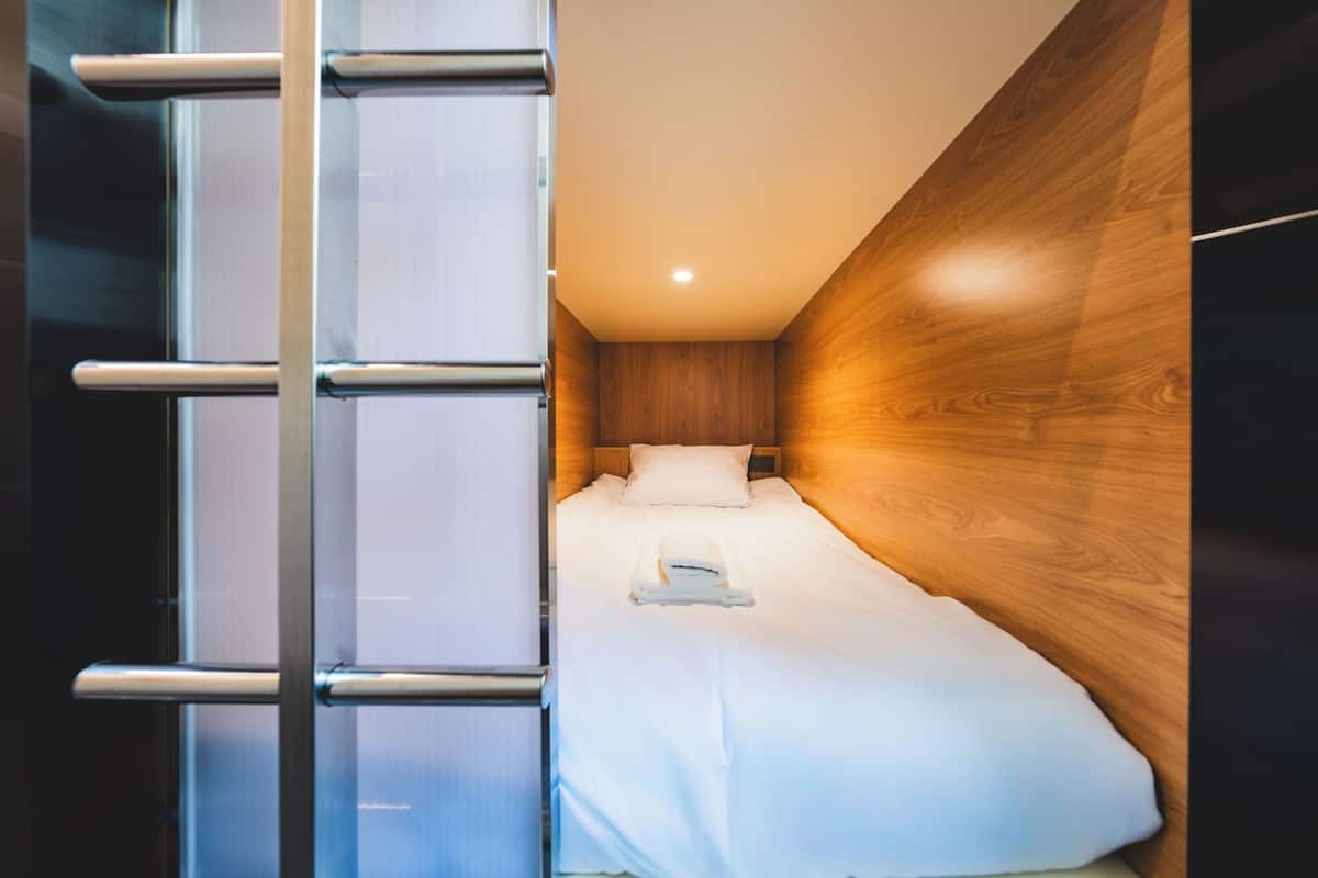 Una habitación pequeña con cama, cabecera de madera, escalera metálica y mesita de noche.